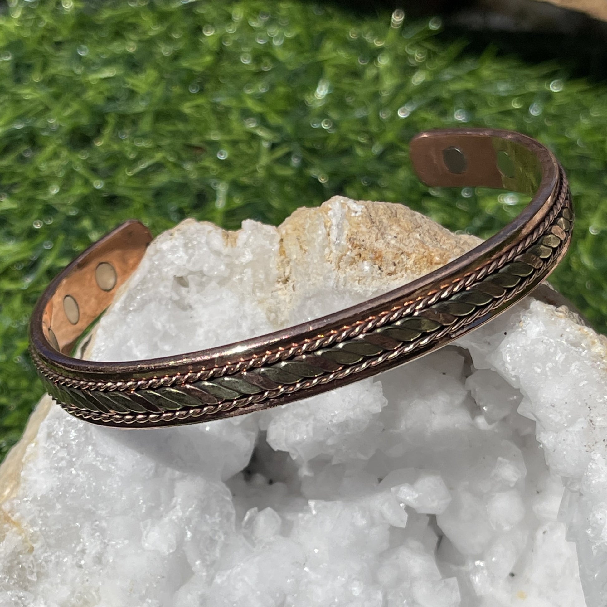 A copper bracelet with a patterned design, displayed on a crystal-like surface.