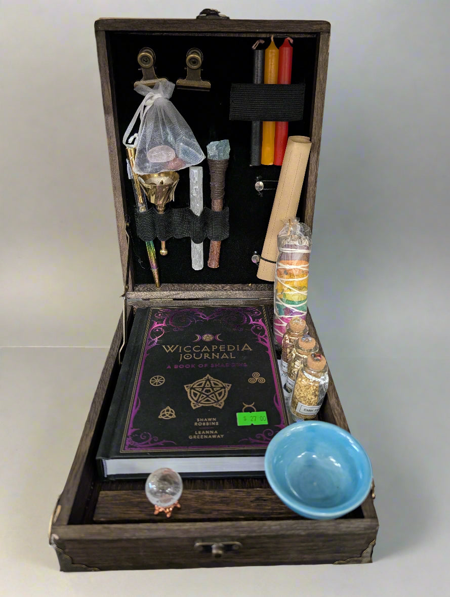 Witchcraft supplies including a Wiccaedia Journal, candles, and tools in an open wooden box.