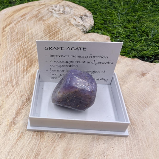 Grape agate stone on a white stand with a card explaining its properties, placed on a wooden surface with grass in the background.